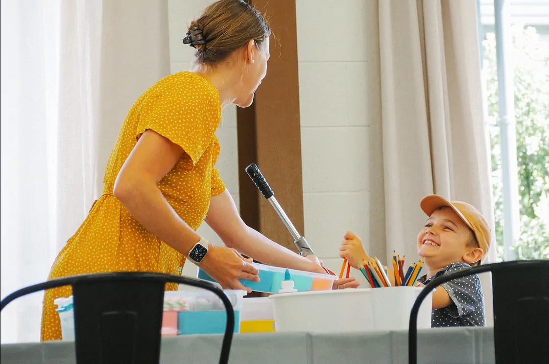 A smiling young boy wearing a beige cap sits at a table filled with art supplies.