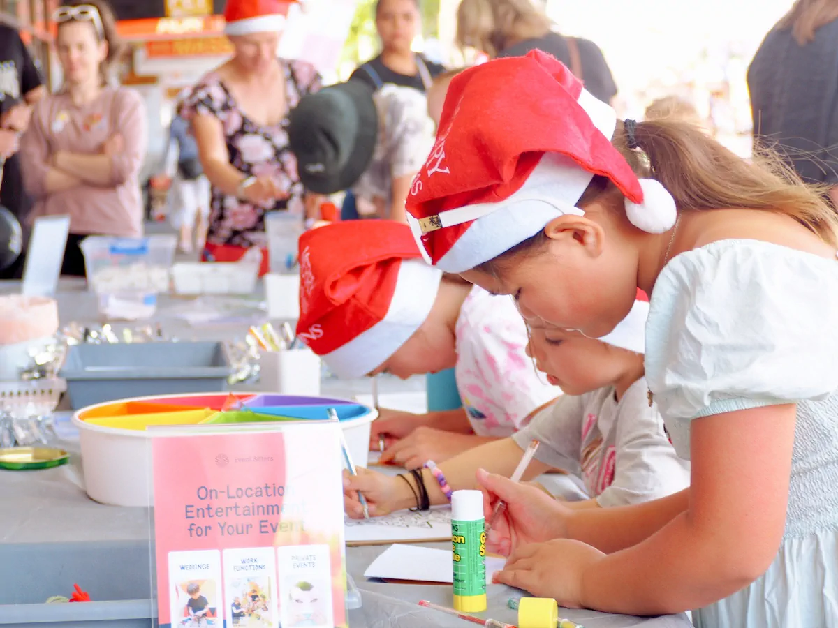 Children in Santa hats doing arts and crafts at an outdoor public event, with Event Sitters signage visible